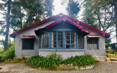 Exterior of a historic grey house with blue window frames and a red roof, surrounded by trees