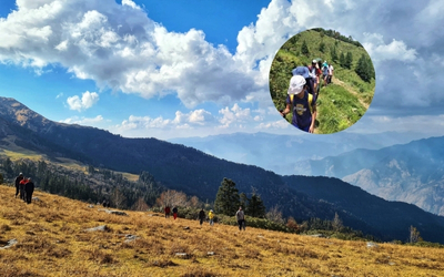 Hikers on a high-altitude meadow with rolling green mountains under a cloudy blue sky